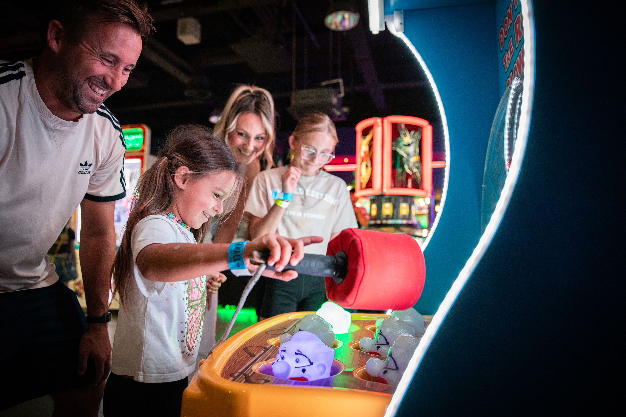 Eine vierköpfige Familie spielt lachend an einem beleuchteten Arcade-Automaten – ein junges Mädchen schlägt mit einem Hammer auf auftauchende Figuren